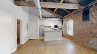 Located at 237-243 Furnace St in Akron, OH, this one-bedroom, one-bathroom loft masterfully blends industrial heritage with contemporary living. The open-concept main area showcases soaring ceilings with exposed black-painted structural rafters, original heavy timber beams, and visible ductwork — all hallmarks of a thoughtfully converted historic building. A striking exposed brick wall runs throughout the space, adding warmth and texture.

The kitchen is well-appointed with stainless steel appliances — including a refrigerator, electric range, microwave, and dishwasher — paired with white upper cabinetry, warm wood lower cabinets, a tile backsplash, and light quartz countertops. A central island provides additional prep and dining space, while pendant lighting adds a modern industrial touch. Light hardwood flooring grounds the main living and kitchen areas.

The bedroom offers a comfortable footprint with plush carpeting, a double-door closet, and the same dramatic exposed brick and original timber beams overhead, with a large arched window allowing natural light to fill the room. The bathroom is clean and modern, featuring a tiled soaking tub and shower combo, wood-toned vanity, quartz countertop, and hardwood flooring — providing a calm, refined retreat within this characterful space.