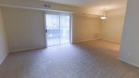 This single-floor residential space offers a clean, functional layout designed for comfortable everyday living. The main living area is generously sized, finished with wall-to-wall neutral carpeting that lends a warm, understated tone throughout. A large sliding glass door fitted with vertical blinds serves as the focal point of the room, flooding the interior with abundant natural light while providing direct access to the outdoors. Glimpses of greenery visible through the glass add a refreshing connection to the outside environment. A pendant light fixture is suspended from the ceiling, offering a subtle decorative touch and supplementary illumination. The walls are finished in a soft, neutral tone, complementing the light and airy atmosphere of the space. Ceiling-mounted air vents and a wall-mounted heating unit indicate a comprehensive climate control system throughout. The overall ambiance is bright, open, and move-in ready, making it a versatile canvas well-suited to a variety of interior styles and furnishing arrangements.