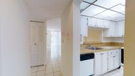This single-floor space offers a clean, functional layout designed for comfortable everyday living. The kitchen is positioned at the heart of the space, featuring white cabinetry with ample upper and lower storage, tile countertops with a warm wood-toned finish, a stainless steel sink, and a built-in dishwasher. A distinctive drop ceiling with a grid panel detail adds visual character overhead. The kitchen opens toward a hallway that connects the main areas of the space, creating an intuitive flow throughout. Light-toned ceramic floor tiles run consistently through the hallway, reflecting natural light and contributing to the overall bright, airy ambiance. A louvered bifold closet door along the hallway provides convenient storage access. Further down the corridor, vertical blinds filter natural light from what appears to be a large window or sliding glass door, illuminating the living area beyond. The walls throughout are finished in a clean, neutral white, giving the space a fresh and open feel. The layout is straightforward and practical, making it easy to navigate while maintaining a cohesive, light-filled atmosphere from the kitchen through to the main living area.