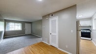 This single-floor space is designed with a practical and straightforward layout. The main living area features a transition between warm hardwood flooring and plush gray carpet, with neutral taupe-gray walls throughout that lend a calm, cohesive ambiance. A louvered closet door and floor vents indicate thoughtful utility integration. Natural light filters in through a window that frames outdoor greenery, enhancing the open feel of the living space.

The kitchen is efficiently arranged with white cabinetry, open upper shelving, dark granite-style countertops, a double stainless steel sink, and a gas range, all set atop hardwood flooring. A window provides additional natural light to the workspace.

The carpeted bedroom is generously proportioned, featuring a large sliding window that overlooks a lush, tree-lined exterior, offering a bright and serene atmosphere. White baseboards frame the room cleanly throughout.

The bathroom is finished with neutral floor tile, a tiled shower-tub surround in a grid pattern, a white vanity with dark countertop, and standard fixtures, providing a clean and functional space. Overall, the unit presents a no-frills, well-maintained interior suitable for comfortable everyday living.