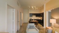 This single-floor space showcases an open-concept design that seamlessly integrates the living, dining, and kitchen areas into one cohesive environment. The living area is anchored by a light blue accent chair with a patterned pillow, offering a welcoming focal point. Elevated ceilings enhance the sense of spaciousness throughout, while track lighting mounted on the ceiling provides directed illumination across the main living space.

The kitchen features dark cabinetry paired with black appliances, including a refrigerator and range, creating a bold contrast against the neutral walls. A functional layout offers ample counter and storage space. Adjacent to the kitchen, a compact dining area accommodates a round wooden table with seating for two, making efficient use of the available space.

Polished concrete-style flooring runs throughout the main area, lending an understated, modern aesthetic. A hallway extending from the living area leads to multiple doors, suggesting additional private rooms such as bedrooms or a bathroom. The overall ambiance is clean, neutral, and functional, with a contemporary feel throughout.