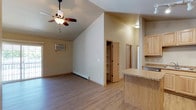 This single-floor residence offers a bright and functional open-concept layout that seamlessly connects the living and kitchen areas. The living space is highlighted by vaulted ceilings that lend a sense of spaciousness and airiness, complemented by a ceiling fan with integrated lighting for year-round comfort. Light-toned hardwood-style flooring runs throughout, adding warmth and continuity to the space. A wall-mounted air conditioning unit and a baseboard heater ensure climate control in every season. The kitchen features light natural wood cabinetry and a granite-topped breakfast bar that serves as both a prep surface and a casual dining area, with stainless steel appliances adding a modern touch. A doorway off the main living area leads to what appears to be a built-in closet or pantry, offering practical storage solutions, while an additional door hints at a bathroom or secondary room beyond. The overall palette of warm neutrals, natural wood tones, and stone surfaces creates a clean, inviting ambiance throughout the home. The layout is thoughtfully designed to maximize both comfort and utility within a well-organized single-level floor plan.