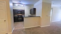 This single-floor space offers a practical and well-organized layout centered around an open-concept kitchen and living area. The kitchen is outfitted with rich, dark wood cabinetry that extends to the upper walls, providing generous storage throughout. Black appliances, including a gas range, microwave, and refrigerator, complement the dark granite-style countertops, creating a cohesive and bold aesthetic. A tiled backsplash adds a refined touch to the cooking area, while the tile flooring in the kitchen transitions smoothly into plush carpeting in the adjoining living space. A peninsula counter with a built-in sink serves as the natural dividing point between the kitchen and living area, offering both additional workspace and a casual bar-style seating option. The living area benefits from the open sightlines created by this layout, giving the space an airy and connected feel. Recessed track lighting illuminates the kitchen, while crown molding along the ceiling adds a subtle architectural detail throughout. Neutral wall tones keep the interior feeling bright and welcoming, providing a versatile backdrop for a range of furnishing styles.