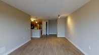 This single-floor residence offers a clean, open-concept design that seamlessly connects the living area to the kitchen. The living space is finished with smooth, light-toned hardwood-style flooring that extends throughout, lending warmth and continuity to the interior. Neutral gray walls provide a versatile backdrop suited to a variety of décor styles, while white baseboards add a crisp finishing touch.

The kitchen is positioned at the far end of the open living area and is equipped with stainless steel appliances, including a full-sized refrigerator. Warm wood-toned cabinetry lines the upper and lower walls, complemented by pendant lighting that hangs above the kitchen counter, creating a welcoming and functional cooking space. A raised breakfast bar provides a casual dining option and helps define the transition between the kitchen and living areas.

A hallway visible from the main living space leads to additional rooms, with a doorway opening to what appears to be a closet or utility space. Track lighting along the hallway ceiling ensures the interior remains well-lit throughout. The overall ambiance is bright, clean, and move-in ready, making this a practical and comfortable single-floor living space.