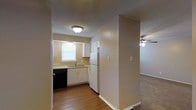This single-story residence showcases a straightforward, functional layout connecting key living areas. The kitchen is equipped with white cabinetry providing ample storage, a double-basin sink positioned beneath a window that allows natural light to filter in, a white refrigerator, and a black dishwasher. The hardwood-style flooring in the kitchen adds warmth and contrast to the neutral beige walls and textured ceiling. A flush-mount ceiling light fixture illuminates the kitchen space effectively. A structural column partially defines the boundary between the kitchen and the adjacent living area, which features carpeted flooring and a ceiling fan, suggesting a comfortable, casual atmosphere. The warm, neutral color palette of beige and white tones is consistent throughout, contributing to a cohesive and understated aesthetic. The open sightlines between the kitchen and living area create a sense of connectivity, making the space feel practical for everyday living. Overall, the residence presents a clean, no-frills interior that prioritizes functionality and ease of movement between its core living spaces.