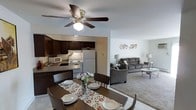 This single-floor residence offers an open-concept layout that seamlessly connects the kitchen, dining area, and living room, creating a cohesive and functional living space. The kitchen is outfitted with dark espresso cabinetry, a white refrigerator, an electric range, a microwave, and a double sink with countertop workspace — providing everything needed for everyday meal preparation. A peninsula-style counter separates the kitchen from the dining area, adding both definition and convenience. The dining area centers around a dark wood table with matching chairs, accented by a decorative table runner and floral centerpiece. A ceiling fan with a light fixture overhead provides both illumination and air circulation. The adjacent living room features plush carpeted flooring and is furnished with a gray sectional sofa, armchairs, a glass-top coffee table, and coordinating floor lamps. The neutral wall tones throughout the space lend a clean, welcoming ambiance. The open flow between all three areas makes the layout ideal for both everyday living and casual entertaining, with each zone maintaining its own sense of purpose while remaining visually connected to the whole.