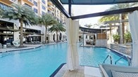 This expansive outdoor pool deck offers a sophisticated resort experience designed for relaxation and leisure. The centerpiece is a large, luminous swimming pool with crystal-clear blue water, framed by clean stone tiling that reflects the open sky above. Elegant white-curtained cabanas line the pool's edge, providing guests with shaded, semi-private retreats that combine comfort with an open-air atmosphere.

Tall, mature palm trees are strategically positioned throughout the space, lending a tropical ambiance and natural shade to the surrounding lounge areas, which are furnished with contemporary chaise lounges and seating. A dedicated poolside bar and lounge area sits to one side, constructed with natural stone and wood accents, offering a stylish setting for refreshments without leaving the pool area.

Overhead pergola structures with retractable shade elements define different zones within the space, adding architectural interest while enhancing guest comfort. The entire area is set against the impressive facade of a multi-story building with a classic, upscale design featuring arched windows and warm-toned finishes. The cohesive design blends modern amenities with a relaxed, sun-drenched aesthetic, making this outdoor pool deck a premier destination for leisure and social gathering.