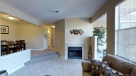 This single-story space offers a comfortable and well-appointed layout with a warm, neutral color palette throughout. The living area serves as the focal point, anchored by a classic tiled fireplace with a black insert, framed by white tile surround and hearth. A decorative wall piece adds an artistic touch above the mantle area, contributing character to the space.

The living area features plush carpeting that defines the seating zone, while tile flooring extends near the fireplace, offering a practical transition between surfaces. A partial wall with a column detail separates the living space from an adjacent area, where a large indoor plant adds a natural, refreshing element.

Steps lead up from the living area, indicating a subtle level change that adds architectural interest to the single-floor layout. A hallway visible in the background connects to additional rooms, with soft overhead lighting illuminating the passage. The corridor hints at a bathroom or utility space beyond.

Overall, the space presents a cozy and functional environment with thoughtful design details, including varied flooring materials, built-in fireplace, and an open yet defined layout that balances comfort with practicality.