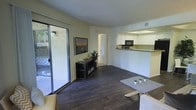 This single-floor space offers a welcoming and functional layout that balances comfort with practicality. The main living area is anchored by rich, dark hardwood flooring that runs throughout, creating a warm and cohesive aesthetic. A large sliding glass door floods the space with natural light and opens directly onto a private patio, offering a seamless indoor-outdoor connection.

The open-concept design allows the living area to flow naturally into the kitchen, which is accessible via a pass-through counter. The kitchen features white cabinetry, black appliances including a built-in microwave, and light-colored countertops, providing a clean, functional workspace. The pass-through design enhances the sense of openness and encourages easy interaction between the two areas.

The living room offers ample space for furnishings, with neutral light-yellow walls that contribute to an airy, relaxed atmosphere. A textured white ceiling adds subtle visual interest overhead. A hallway visible from the main living area suggests additional private rooms beyond, completing the practical layout. Overall, the space presents a bright, well-connected environment suited for comfortable everyday living.