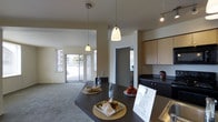 This single-floor space is designed with an open-concept layout that seamlessly connects the kitchen and living area, creating a cohesive and functional environment. The kitchen is anchored by a long, dark-topped island or peninsula that serves as both a prep and gathering surface, complemented by light wood cabinetry above and dark lower cabinetry that provides ample storage. A series of pendant lights hang at varying heights above the kitchen workspace, adding a contemporary touch, while a track light fixture near the ceiling provides additional illumination. The countertops carry a dark tone that contrasts cleanly with the neutral wall colors throughout. The living area beyond features carpeted flooring and is bathed in natural light courtesy of large sliding glass doors fitted with vertical blinds, offering a view to an outdoor space. The overall color palette is understated and neutral, with warm beige tones on the walls and ceilings contributing to a calm, welcoming ambiance. A doorway visible in the background hints at additional rooms beyond the main open living space, suggesting a practical and well-organized floor plan throughout.