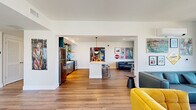 Located at 2121 Wood St in Oakland, CA, this single-floor communal space is a vivid showcase of contemporary design and urban creativity. The open-plan layout seamlessly connects the kitchen and living area, creating a generous, free-flowing environment bathed in natural light from large windows that frame views of the surrounding Oakland cityscape.

The kitchen is a standout feature, equipped with stainless steel appliances — including a full-size refrigerator, range, and dishwasher — paired with sleek teal cabinetry and a white countertop island with bar seating. The most striking element is the bold, graffiti-inspired mural that serves as a backsplash, infusing the kitchen with an unmistakable artistic energy.

The living area is divided into distinct seating zones: a lounge corner with deep navy blue walls, leather sectional sofas, and café-style tables positioned beneath bright windows, and a more casual area featuring a mustard yellow accent chair, wall-mounted TV, and vibrant artwork throughout.

The hallway entry welcomes visitors with warm wood-look flooring, colorful large-scale art pieces on crisp white walls, and a relaxed seating arrangement with bean bag chairs near the glass double doors. Recessed lighting and clean architectural lines maintain a polished yet approachable atmosphere across the entire space.
