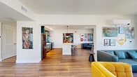 Located at 2121 Wood St in Oakland, CA, this single-floor communal space is a vivid showcase of contemporary design and urban creativity. The open-plan layout seamlessly connects the kitchen and living area, creating a generous, free-flowing environment bathed in natural light from large windows that frame views of the surrounding Oakland cityscape.

The kitchen is a standout feature, equipped with stainless steel appliances — including a full-size refrigerator, range, and dishwasher — paired with sleek teal cabinetry and a white countertop island with bar seating. The most striking element is the bold, graffiti-inspired mural that serves as a backsplash, infusing the kitchen with an unmistakable artistic energy.

The living area is divided into distinct seating zones: a lounge corner with deep navy blue walls, leather sectional sofas, and café-style tables positioned beneath bright windows, and a more casual area featuring a mustard yellow accent chair, wall-mounted TV, and vibrant artwork throughout.

The hallway entry welcomes visitors with warm wood-look flooring, colorful large-scale art pieces on crisp white walls, and a relaxed seating arrangement with bean bag chairs near the glass double doors. Recessed lighting and clean architectural lines maintain a polished yet approachable atmosphere across the entire space.