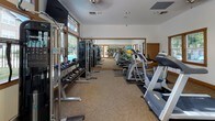 This fitness center offers a comprehensive workout environment designed to accommodate a wide range of exercise needs. The space is thoughtfully laid out across a single floor, with durable, textured carpeted flooring throughout and clean white walls accented by warm wood trim details that lend a polished, welcoming feel.

The cardio zone features multiple treadmills and an elliptical machine arranged in a row, complemented by large floor-to-ceiling mirrors that visually expand the space and allow users to monitor their form. Natural light streams in through several wood-framed windows positioned along the walls and at elevated heights, providing bright, airy conditions throughout the day. A wall-mounted flat-screen television adds an entertainment option for cardio sessions.

The strength training area is well-appointed with a Precor functional training cable machine, a multi-station cable tower, and a full rack of dumbbells ranging across various weights. A weight bench provides additional versatility for resistance training. A dedicated accessory station holds an assortment of stability balls, resistance bands, and balance tools, supporting functional and rehabilitative fitness routines. A McKenzie exercise information board is posted on the wall, hinting at a focus on structured, health-oriented programming.

Glass-paned double doors at the rear of the space open to an adjacent area, enhancing the sense of openness and connectivity within the facility.