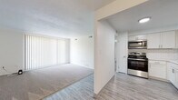 This single-floor residence offers a clean, modern aesthetic throughout its well-organized layout. The open-concept living and dining area is anchored by light wood-look vinyl flooring that flows seamlessly from the kitchen into the main living space, transitioning to plush wall-to-wall carpeting in the living room. A ceiling fan with a light fixture adds both function and a finishing touch to the space. Large vertical blinds along the living room window allow for adjustable natural light, creating a bright and airy atmosphere.

The kitchen has been thoughtfully updated with crisp white cabinetry, sleek white quartz countertops, and a classic white subway tile backsplash. A full suite of stainless steel appliances — including a refrigerator, range, over-the-range microwave, and a stainless steel sink — rounds out this functional and stylish cooking space.

A hallway with wood-look flooring connects the main living areas to the additional rooms, with a closet providing convenient storage. The bathroom features large-format stone-look floor tiles, a fully tiled tub-and-shower surround in a neutral taupe palette, a white vanity, and brushed nickel fixtures, conveying a spa-like sense of calm. Throughout, the space is finished in bright white walls, lending a fresh, move-in-ready feel.