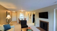 This single-floor residence is designed with an open-concept layout that seamlessly connects the living, dining, and kitchen areas, creating a cohesive and functional living environment. The living room serves as the central gathering space, highlighted by a tile-surround fireplace that anchors the room and adds warmth. A large flat-screen television is mounted on the wall above the fireplace, contributing to the room's entertainment-ready setup. The space is finished with wall-to-wall carpet in a neutral tone, complementing the light beige walls throughout.

Adjacent to the living area, the dining space accommodates a glass-top table with red-upholstered chairs, positioned near a window that allows natural light to filter in. The open sightline to the kitchen, which features dark cabinetry and black appliances, ensures ease of movement and interaction between areas. Flush-mount ceiling fixtures provide consistent overhead lighting throughout. A textured popcorn ceiling is present throughout the main living space. Doorways lead to additional rooms, suggesting a practical floor plan with clear separation between living and private spaces. The overall ambiance is casual and comfortable, with a neutral palette suited to a variety of decor styles.