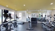 This fitness center offers a clean, modern environment designed to support a full range of workout routines. The space is defined by its striking yellow ceiling, which adds a vibrant energy to the room, while recessed lighting ensures the entire area is well-illuminated throughout the day. Large windows along one wall allow natural light to filter in, creating an open and inviting atmosphere.

The main workout floor is finished with durable dark rubber flooring, providing both safety and comfort during exercise. A full wall of mirrors runs along one side of the space, visually expanding the room and allowing users to monitor their form while training.

Cardio equipment includes treadmills, a stationary bike, and a rowing machine, offering a variety of aerobic training options. The free weight section features a well-stocked dumbbell rack with a wide range of weights neatly organized for easy access. Strength and functional training equipment, including a cable machine and stability balls, round out the facility's offerings.

Decorative fitness-themed artwork on the walls contributes to the motivating aesthetic of the space. The layout is thoughtfully arranged to provide clear pathways between equipment, ensuring a safe and comfortable experience for all users.