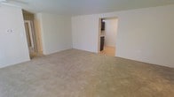 This single-floor residential space offers a clean and straightforward layout designed for comfortable everyday living. The main living area is generously sized, featuring wall-to-wall neutral carpeting that adds warmth underfoot and a sense of openness throughout the room. The walls are finished in a crisp, neutral tone, contributing to a bright and airy atmosphere that makes the space feel inviting and easy to personalize.

A wide open passageway connects the living area to the kitchen, creating a natural flow between the two spaces. The kitchen features hardwood-style flooring that contrasts with the carpeted living area, along with dark cabinetry that provides a bold, functional aesthetic. The transition between the two rooms is seamless, making the overall layout feel cohesive and practical.

The space is finished simply and cleanly, with minimal architectural ornamentation, lending it a versatile character suited to a variety of interior styles. The neutral color palette throughout creates a calm, understated ambiance that serves as an excellent backdrop for furnishing and décor. A doorway on one side of the living area suggests additional rooms or storage, further enhancing the functional appeal of this well-organized single-floor layout.