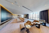 Located at 257 NE 29th Street in Miami's vibrant Edgewater district, this elegantly appointed lounge occupies a single expansive floor with a generously open layout. The space is defined by a wall of floor-to-ceiling windows along one side, flooding the interior with abundant natural light and offering panoramic views of the Miami skyline. Smooth white ceilings with recessed and track lighting complement warm ambient cove lighting that runs along a richly paneled feature wall, creating a refined and inviting atmosphere throughout. A large built-in display wall anchored by dark wood-toned cabinetry serves as a commanding focal point, ideal for presentations or entertainment. The interior is furnished with plush, neutral-toned sectional sofas, cognac leather lounge chairs, and sleek round accent tables, all arranged to encourage both relaxed conversation and formal gathering. A striped carpet in warm beige tones grounds the space and adds subtle texture underfoot. A secondary seating vignette toward the rear features colorful curated artwork displayed on a fluted accent wall, adding a creative and lively touch. Lush indoor greenery contributes a fresh, organic element to the contemporary design. The overall ambiance is polished and modern, well-suited for upscale social or professional use.