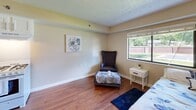 This single-story unit at King's Reserve College Park in Indianapolis offers an efficiently designed layout across one floor. The generously sized primary bedroom occupies the majority of the floor plan, featuring warm hardwood-style flooring, a textured ceiling, and neutral beige walls that create a calm, inviting atmosphere. A large window allows natural light to fill the room, offering an outdoor view of the surrounding grounds. The bedroom provides ample space for a full seating area in addition to standard furnishings.

The kitchen is well-appointed with white cabinetry extending to the ceiling for maximum storage, a gas range, full-size refrigerator, and a stainless steel sink with a dark countertop. Wood-style flooring continues throughout, maintaining a cohesive look. An entrance hallway connects the main spaces seamlessly.

The bathroom features warm hardwood-style flooring, a rich dark-wood vanity with a white countertop, well-lit mirror with overhead vanity lighting, and a shower-tub combination enclosed by a decorative blue floral curtain. Clean white walls and coordinated blue accents give the bathroom a tidy, polished appearance throughout.