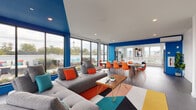 This single-floor space makes a striking first impression with its bold cobalt blue accent walls and ceilings that contrast sharply against crisp white surfaces throughout. The open-plan layout connects a generous lounge area, a dining zone, and a fully equipped kitchen in one cohesive, flowing environment. Floor-to-ceiling windows wrap around the lounge and dining areas, flooding the space with abundant natural light and offering expansive views of the outdoor terrace, which features additional seating and greenery. The lounge is anchored by a large sectional sofa accented with teal and orange throw pillows, arranged around a mid-century modern wood coffee table atop a bold geometric area rug in teal, orange, yellow, and charcoal. A wall-mounted flat-screen television adds functionality to the living area. The dining zone accommodates a long communal table surrounded by vibrant orange chairs, illuminated by pendant lighting. The kitchen is sleek and modern, featuring white cabinetry extending to the ceiling, stainless steel appliances including a range and French door refrigerator, a white quartz island with bar seating, dark hardwood flooring, and white subway tile backsplash. Throughout, the design blends energetic color with contemporary minimalism, creating a dynamic yet functional shared environment.