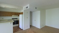 This single-story space offers a practical, open-concept layout where the kitchen and living area flow seamlessly into one another. The kitchen is equipped with essential appliances, including a black electric range and a white refrigerator, complemented by warm wood-toned cabinetry that provides ample storage. An under-cabinet light fixture adds functional illumination to the cooking area, while a range hood sits above the stove for ventilation.

A textured popcorn ceiling runs throughout, adding a classic character to the space. The flooring transitions from hard surface wood-look material in the kitchen to carpeting in the living area, delineating the two zones naturally. A central structural column divides the space and houses HVAC vents, ensuring climate control throughout.

The living area is open and simply finished, with neutral white walls and clean baseboards that lend a bright, airy feel. A doorway visible beyond the column suggests access to additional areas of the unit. Overall, the space presents a straightforward, functional design suited to comfortable everyday living.