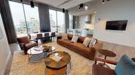 This elegantly designed single-floor space functions as a high-end communal lounge or amenity suite with a distinctly modern urban character. The open-plan layout flows seamlessly across multiple seating and gathering areas, all unified by warm hardwood flooring and a cohesive contemporary aesthetic.

The main lounge area is anchored by a striking marble-clad fireplace wall, which also frames a wall-mounted flat-screen television. Rich cognac leather sofas, velvet accent chairs, and layered area rugs add warmth and texture throughout. Multiple seating vignettes encourage both intimate conversation and larger group gatherings.

Floor-to-ceiling windows wrap the perimeter, flooding the space with natural light and offering sweeping views of the surrounding cityscape. A separate lounge alcove with direct access to an outdoor terrace provides additional seating with panoramic urban views.

The fully equipped kitchen features sleek white cabinetry, a dramatic marble slab backsplash, a stainless steel range hood, a gas cooktop, and a central island with bar seating illuminated by pendant lighting. An adjacent dining area accommodates a large group table, making the space well-suited for events and entertaining. The overall ambiance is polished, sophisticated, and inviting.