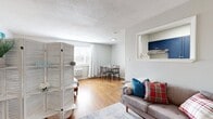 This single-floor studio is thoughtfully laid out to maximize functionality within a compact footprint. The main living area features warm hardwood flooring, soft neutral walls, and a textured ceiling that lends the space a cozy, finished feel. Natural light filters in through a window dressed with white curtains, brightening the open-plan living and dining zone, where a loveseat and small dining set create distinct yet connected areas. A white louvered room divider elegantly separates the sleeping zone, which comfortably accommodates a full-sized bed, nightstands, and a media console. A pass-through window connects the living area to the galley-style kitchen, which features bold navy shaker cabinets, stainless steel appliances including a full-sized refrigerator and range, and light hardwood-style flooring. The kitchen is narrow but efficiently organized with ample cabinetry. The bathroom is well-appointed with white subway tile wainscoting, patterned floor tile, a dark wood vanity with chrome fixtures, and a mirrored medicine cabinet. Two hallways provide access between the space's functional zones, contributing to an efficient and organized overall flow throughout the unit.
