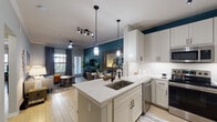 This thoughtfully designed single-floor residence showcases a cohesive modern aesthetic anchored by a bold teal and neutral color palette throughout. The open-concept layout seamlessly connects the kitchen and living area, creating a fluid and functional shared space. The kitchen is outfitted with crisp white shaker cabinetry extending to the ceiling, quartz countertops, stainless steel appliances including a range, built-in microwave, and dishwasher, and a central peninsula with an undermount sink and stylish pendant lighting overhead. The living area features crown molding, light wood-look tile flooring, a ceiling fan, and large windows with draping curtains that invite generous natural light and outdoor views. The primary bedroom is a serene retreat with a bay window configuration, a deep teal accent wall, and direct access to a walk-in closet. A second bedroom offers similar design elements, including a matching accent wall and walk-in closet access. The bathroom presents a full soaking tub with marble-look tile surround, a single vanity with white cabinetry and quartz countertop, and matte black fixtures that add a refined, contemporary contrast. Throughout the residence, consistent finishes and intentional design details contribute to a polished and inviting ambiance.
