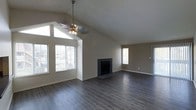 This single-floor home offers an inviting and well-designed living space that balances architectural character with functional layout. The main living area is the centerpiece of the home, featuring dramatic vaulted ceilings that lend a sense of openness and volume to the room. A stylish ceiling fan with a multi-bulb light fixture hangs as a focal point, complementing the angled roofline above.

Dark hardwood-style flooring runs throughout the space, providing a rich, grounded contrast to the neutral warm-gray wall tones. A classic fireplace with a dark surround anchors one wall of the living area, offering both aesthetic appeal and practical warmth. Large, multi-pane windows flood the space with generous natural light, framing views of the surrounding exterior and creating a bright, airy atmosphere throughout the day.

The open floor plan connects the main living area to an additional space, potentially suited for a dining area or secondary seating arrangement, with its own window providing continued natural light. The clean, neutral palette throughout the home provides a versatile backdrop, making it easy to personalize. Overall, this home combines architectural interest with a warm, livable ambiance.