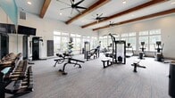 Located at 175 Main Street Market Place SE in Cartersville, Georgia, this well-appointed fitness center offers a generously sized, single-floor exercise space designed to support a comprehensive workout experience. The room's architecture is defined by soaring ceilings adorned with distinctive exposed wood beams and recessed lighting, creating an open and energizing atmosphere. Large, multi-pane windows line the walls, flooding the interior with abundant natural light and offering views of the surrounding greenery. Ceiling fans are strategically placed throughout to ensure comfortable airflow during workouts. The flooring features patterned, multi-tone carpet with teal accents that complement the room's bold teal accent wall, which serves as a vibrant backdrop to a full-length wall mirror — ideal for monitoring form and making the space feel even larger. The fitness center is thoroughly equipped with a wide array of cardio machines, including treadmills, ellipticals, a stair climber, and stationary bikes, as well as multi-station strength training machines, adjustable benches, and a well-stocked dumbbell rack. The thoughtful layout provides ample floor space between equipment, allowing for comfortable movement and flexibility training throughout the room.