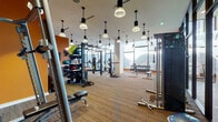 This fitness center is a thoughtfully designed, single-level space that combines functionality with a contemporary aesthetic. Expansive floor-to-ceiling windows line the perimeter, flooding the interior with natural light and offering sweeping panoramic views of the surrounding landscape—an energizing backdrop for any workout.

The space is divided into distinct functional zones. A dedicated cardio area houses multiple treadmills and a curved manual treadmill, stationary bikes, and rowing machines, all positioned to take full advantage of the scenic outdoor views. An open strength training area features adjustable benches, a well-stocked dumbbell rack, a power rack with cable attachments, and battle ropes, providing comprehensive options for resistance training.

A standout feature is the climbing wall, branded as a "Freedom Climber," which adds a unique and dynamic element to the fitness offering. Warm terracotta-orange accent walls contrast with neutral carpet flooring and white structural columns, creating an inviting yet motivating atmosphere.

Pendant lighting throughout adds a polished, modern touch, while a built-in refreshment station—complete with a mini-fridge, storage cabinetry, and a wall-mounted TV—offers added convenience and comfort for users.