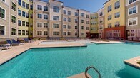 Located at 630 Cotanche St in Greenville, North Carolina, this outdoor amenity space is the centerpiece of a multi-story residential community. The courtyard is enclosed by multi-story buildings featuring a mix of taupe horizontal lap siding, brick accents, and large multi-pane windows that provide an attractive architectural backdrop to the outdoor environment.

The highlight of the space is a generously sized resort-style swimming pool with clear, blue water, framed by an expansive concrete sun deck. A row of blue lounge chairs lines the deck, offering ample space for relaxation in the open air. A pergola structure near the pool adds a shaded gathering area, enhancing the recreational appeal of the space.

Controlled access to the courtyard is provided through a secure, gated entry featuring wrought iron-style fencing and glass panel doors, ensuring a private and safe environment. Raised landscaped planters with mulched beds and seasonal greenery are positioned along the building perimeters, softening the hardscape and adding natural texture throughout. Floor-to-ceiling glass walls on portions of the building exterior allow interior amenity spaces to look out directly onto the pool area. Perimeter walkways connect all areas of the courtyard, providing convenient and accessible circulation throughout this well-maintained outdoor amenity space.