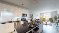 This thoughtfully designed single-floor residence combines contemporary finishes with a functional open layout. The kitchen serves as a focal point, showcasing high-gloss white cabinetry, dark granite countertops, a subway tile backsplash, and a full suite of stainless steel appliances including a refrigerator, range, microwave, and dishwasher. A central island with seating provides additional prep space and a casual dining option. Track lighting overhead enhances the kitchen's clean, modern aesthetic.

The kitchen flows seamlessly into the living area, which features warm wood-look flooring, neutral walls, and a door leading to a private outdoor space. Large windows fill the living area with natural light and offer pleasant outdoor views, creating a bright and inviting atmosphere throughout.

The bedroom features a striking blue accent wall, a large window with scenic views, a dedicated walk-in closet with shelving, and direct access to an en-suite bathroom. The bathroom includes a wood-toned vanity with a white countertop, tile flooring, and a tub-shower combination. A stacked washer and dryer unit is conveniently located adjacent to the bathroom, adding practical everyday functionality to the residence.