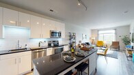 This thoughtfully designed single-floor residence combines contemporary finishes with a functional open layout. The kitchen serves as a focal point, showcasing high-gloss white cabinetry, dark granite countertops, a subway tile backsplash, and a full suite of stainless steel appliances including a refrigerator, range, microwave, and dishwasher. A central island with seating provides additional prep space and a casual dining option. Track lighting overhead enhances the kitchen's clean, modern aesthetic.

The kitchen flows seamlessly into the living area, which features warm wood-look flooring, neutral walls, and a door leading to a private outdoor space. Large windows fill the living area with natural light and offer pleasant outdoor views, creating a bright and inviting atmosphere throughout.

The bedroom features a striking blue accent wall, a large window with scenic views, a dedicated walk-in closet with shelving, and direct access to an en-suite bathroom. The bathroom includes a wood-toned vanity with a white countertop, tile flooring, and a tub-shower combination. A stacked washer and dryer unit is conveniently located adjacent to the bathroom, adding practical everyday functionality to the residence.