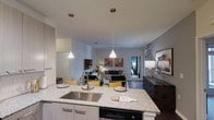Located at 201 S Elliott Rd in Chapel Hill, NC, this single-floor residence offers a thoughtfully designed open-concept layout that seamlessly connects the kitchen, dining, and living spaces. The kitchen serves as a central hub, anchored by a generously sized granite island with a stainless steel undermount sink, crisp white cabinetry with sleek hardware, and an integrated dishwasher. Stylish cone-shaped pendant lights hang above the island, providing focused illumination while adding a contemporary aesthetic touch. The dining area transitions naturally into the living room, where a comfortable seating arrangement creates an inviting atmosphere for both relaxing and entertaining. The living space is accented with warm décor, including a neutral sofa, mustard-toned accent chairs, and curated wall art that adds a pop of color. Light-toned wood-style flooring runs throughout, contributing to a clean, modern look. A doorway visible from the main living area leads to a private bedroom beyond, offering a sense of depth and separation between living and sleeping spaces. The overall ambiance is contemporary and welcoming, with a cohesive design palette of neutral tones, warm accents, and modern fixtures throughout.