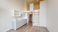 Located at 2211 SW Fourth Ave in Portland, Oregon, this efficiently designed unit makes exceptional use of vertical space through a dramatic loft configuration. Soaring ceilings define the main living and kitchen area, where a built-in loft platform is accessed via a wooden ladder, creating a dedicated sleeping space above without sacrificing square footage below. The kitchen is outfitted with gray cabinetry, white countertops, a stainless steel sink, cooktop, microwave, and refrigerator, along with open wall-mounted shelving for added storage. Wood-look plank flooring runs throughout, lending warmth and continuity across the space. Oversized windows, including floor-to-ceiling panels and a glass door that opens to a small balcony, flood the living area with natural light and offer views of the surrounding Portland cityscape and greenery. A ceiling fan adds comfort to the airy main room. The hallway connects to a bathroom featuring a walk-in shower, toilet, and vanity, all finished in clean white tones with wood-look tile flooring and stainless steel grab bars. A closet with wire shelving provides practical storage just off the hallway. The overall aesthetic is clean, modern, and minimalist.