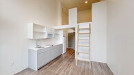Located at 2211 SW Fourth Ave in Portland, Oregon, this efficiently designed unit makes exceptional use of vertical space through a dramatic loft configuration. Soaring ceilings define the main living and kitchen area, where a built-in loft platform is accessed via a wooden ladder, creating a dedicated sleeping space above without sacrificing square footage below. The kitchen is outfitted with gray cabinetry, white countertops, a stainless steel sink, cooktop, microwave, and refrigerator, along with open wall-mounted shelving for added storage. Wood-look plank flooring runs throughout, lending warmth and continuity across the space. Oversized windows, including floor-to-ceiling panels and a glass door that opens to a small balcony, flood the living area with natural light and offer views of the surrounding Portland cityscape and greenery. A ceiling fan adds comfort to the airy main room. The hallway connects to a bathroom featuring a walk-in shower, toilet, and vanity, all finished in clean white tones with wood-look tile flooring and stainless steel grab bars. A closet with wire shelving provides practical storage just off the hallway. The overall aesthetic is clean, modern, and minimalist.