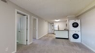 Located at 133 Belchertown Rd in Amherst, Massachusetts, this single-floor unit offers a practical and well-organized layout with a clean, neutral aesthetic throughout. The open living area is finished with plush carpeting and flows directly into a kitchen and utility zone. The kitchen is appointed with white shaker-style cabinetry, dark stone-look countertops, light wood-look flooring, and a full suite of stainless steel appliances including a range, microwave, and dishwasher. A stacked in-unit washer and dryer is conveniently positioned adjacent to the kitchen, adding everyday functionality. The living space connects to sliding glass doors that open to a lush outdoor view, filling the room with natural light. Multiple bedrooms are carpeted and well-proportioned, each featuring windows with vertical blinds that provide both privacy and access to natural light. One bedroom includes a sliding closet door for efficient storage. The unit includes two bathrooms: one features a full tub-shower combination with clean white fixtures, while the second is appointed with a white shaker vanity, dark countertop, large mirror, and wood-look tile flooring. The textured ceilings and neutral wall tones carry throughout, creating a cohesive and move-in ready environment.