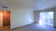 This single-floor residential space offers a clean and functional layout with a straightforward design. The living area features wall-to-wall carpeting in a neutral tone, providing a comfortable and cohesive foundation throughout the space. The walls are finished in a light, neutral color palette that contributes to a bright and open atmosphere. A textured popcorn ceiling adds a classic residential touch overhead. Natural light is a notable feature, streaming in through a sliding glass door fitted with vertical blinds, which offers both privacy control and access to the outdoors. The sliding door serves as the primary source of natural illumination, casting light across the carpeted floor and enhancing the airy feel of the room. The overall design is minimalist and move-in ready, offering a blank canvas for personalization. The space is well-suited for everyday living, with its simple finishes and practical layout making it an accessible and functional environment. The single-floor configuration ensures ease of movement throughout, making the space comfortable and straightforward to navigate.