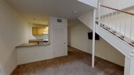 Located at 1375 W Broad St in Richmond, Virginia, this two-story residence offers a practical and functional layout spread across two levels. The main living area is carpeted and open, anchored by a white staircase with a railing that leads to the upper floor. A support column near the staircase adds a structural architectural element to the space. A wall-mounted TV bracket is positioned beneath the staircase, making efficient use of the available wall space. A louvered interior door provides access to a closet or utility area, contributing to built-in storage options. Adjacent to the living area, the kitchen features light wood cabinetry, a countertop bar that opens toward the living space, a range, and a sink. The kitchen is finished with tile flooring that continues into the adjacent utility or laundry area, which accommodates additional appliances. The bathroom is clean and simply appointed, featuring white tile flooring, a full-size soaking tub with a shower and curtain rod, a toilet, and a wood-toned vanity with a granite-style countertop. Chrome hardware and a towel bar add functional finishing touches. The overall interior presents a neutral color palette of whites and beiges, offering a straightforward and move-in ready living environment.