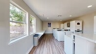 This shared laundry facility is a clean, efficiently designed space laid out on a single floor. The room is finished with light wood-look luxury vinyl plank flooring and neutral-toned walls, creating a bright and welcoming environment. Recessed lighting throughout the ceiling supplements the generous natural light provided by multiple large windows, which offer views of the surrounding outdoor area.

The facility is equipped with a full lineup of commercial-grade top-loading washers and stacked washer-dryer units, all in white, arranged to maximize usable floor space. Built-in white folding counters run along the walls in multiple areas, providing ample surface area for sorting and folding laundry. Waste bins are conveniently positioned near the counters for ease of use.

Ventilation is handled through ceiling-mounted HVAC vents, ensuring a comfortable environment year-round. A fire extinguisher is visibly mounted on the wall, reflecting attention to safety standards. Informational signage and posted instructions are displayed for user reference. The overall layout is open and functional, designed to accommodate multiple users simultaneously while maintaining a tidy, organized atmosphere.