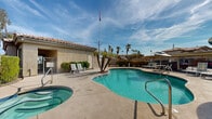 Located in Phoenix, Arizona, this single-level outdoor amenity area offers a well-appointed communal retreat designed for relaxation and recreation in the heart of the Sonoran Desert. The centerpiece is a generously sized freeform swimming pool with vivid turquoise water and clearly marked depth indicators, complemented by a separate circular spa/hot tub positioned nearby for easy access. The surrounding concrete sun deck provides ample space for lounging, furnished with multiple sets of patio chairs, chaise lounges, and shaded umbrella tables, creating comfortable spots for both sunbathing and socializing. A compact utility structure with a tile roof is situated at the edge of the deck, consistent with the Spanish-influenced architectural style seen throughout the surrounding buildings, which feature matching stucco exteriors and clay tile rooflines. The area is enclosed by a combination of iron fencing, block walls, and dense hedging, offering privacy while maintaining an open, airy feel. Mature palm trees and native landscaping add a distinctly Southwestern character to the space. Under the broad, sun-filled Phoenix sky, this outdoor area delivers a resort-like atmosphere ideal for enjoying the region's warm climate year-round.
