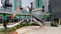 This dynamic outdoor courtyard is part of the Access Culver City development, a contemporary mixed-use property distinguished by its bold architectural design. The building exterior showcases a modern aesthetic with eye-catching teal/green corrugated metal cladding, dark grey paneling, and glass-railed balconies that overlook the ground-level plaza below.

At the heart of the courtyard stands a commanding large-scale stainless steel sculpture, its reflective, sweeping ribbon-like form serving as a dramatic focal point for the space. The polished metalwork contrasts beautifully against the surrounding architecture and landscaping.

The ground level features smooth concrete walkways and plazas, accented by curved concrete planters filled with lush, drought-tolerant plantings including striking echium, ornamental grasses, and a variety of low-water succulents and flowering plants. Warm-toned wooden benches and classic black street lamps are thoughtfully placed throughout, providing both comfort and ambient lighting for visitors.

A glass-and-steel staircase connects the ground-level courtyard to an upper terrace, adding vertical dimension and accessibility to the space. The overall ambiance is modern, urban, and welcoming — designed to serve as a lively community hub that blends art, nature, and contemporary architecture seamlessly.