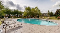 Located on Hannah Ave in Norristown, PA, this outdoor pool area is a well-appointed community amenity designed for relaxation and recreation. The centerpiece is a generously sized oval swimming pool with clear, crystal-blue water, offering ample space for both leisurely swimming and casual enjoyment. The pool features step-entry access points, making it accessible and inviting.

Surrounding the pool is a broad, paved stone deck finished in neutral tones, providing plenty of room for circulation and lounging. Multiple adjustable chaise lounge chairs are arranged around the perimeter, offering comfortable spots to unwind and soak in the open-air setting. Manicured green lawn areas border the deck, adding a natural softness to the hardscape.

The space is enclosed by a classic black iron perimeter fence, providing both security and an unobstructed view of the surrounding mature trees and landscaping. Tall deciduous trees frame the area on multiple sides, offering partial shade and a sense of seclusion while maintaining an open, airy atmosphere under an expansive sky. Decorative potted plantings are thoughtfully placed throughout the space, adding greenery and visual interest.

The overall ambiance is clean, well-maintained, and community-oriented, making this outdoor pool area an appealing retreat within the Norristown residential neighborhood.