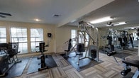 This single-floor fitness center is designed to support a comprehensive workout experience. The space is furnished with a notable selection of strength-training equipment, including a multi-station cable machine, a dedicated weight bench, and additional resistance machines positioned throughout the room. Rubber exercise mats are placed beneath the equipment to provide stability and floor protection. The flooring features a neutral, striped carpet tile that complements the light beige walls, creating a clean and functional environment. Natural light enters through multiple windows along the perimeter walls, lending the space an open and airy feel, while recessed and overhead fluorescent lighting ensures consistent illumination throughout. A textured ceiling adds a subtle architectural detail to the room. The layout is thoughtfully organized, allowing users to move between stations with ease. Wall-mounted accessories, such as a paper towel or wipe dispenser, reflect attention to hygiene and convenience. Overall, the fitness center provides a practical and well-maintained setting suitable for a range of strength and conditioning routines.