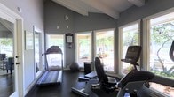 This single-floor fitness center offers a welcoming and functional workout environment designed to keep residents active and motivated. The space is characterized by soaring vaulted ceilings with exposed beam detailing, lending an open and airy feel that enhances the overall experience. Large white-framed windows line the walls on multiple sides, flooding the interior with abundant natural light and providing pleasant views of the surrounding outdoor amenity area, which features mature palm trees, manicured landscaping, and patio seating. The flooring is a durable dark rubber material, ideal for a fitness setting. Equipment includes a treadmill, a recumbent exercise bike, and a stability ball, offering a variety of cardiovascular and core training options. The walls are finished in a clean, neutral gray tone that creates a calm and focused atmosphere. A posted rules and guidelines board near the entrance keeps the space organized and user-friendly. The combination of natural light, high ceilings, and tropical outdoor scenery visible through the windows makes this fitness center a particularly inviting space for daily exercise routines.