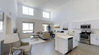 This contemporary two-story space makes a striking first impression with its soaring double-height ceilings that flood the open-concept main level with an abundance of natural light. Oversized windows, including clerestory windows positioned high on the walls, amplify the sense of volume and airiness throughout the living area.

The main floor is designed around a seamless open-concept layout that integrates the kitchen, dining, and living spaces. The modern kitchen features crisp white cabinetry, a clean-lined peninsula with a built-in sink and stainless steel dishwasher, and light countertops that contribute to the bright, airy aesthetic. The kitchen flows effortlessly into the dining area and an expansive living room, making the space well-suited for both everyday living and entertaining.

The living area is anchored by light-toned hardwood-style flooring that runs throughout, complementing the neutral white walls and contemporary furnishings. A sliding glass door provides access to an outdoor area, further enhancing the connection between interior and exterior. The overall ambiance is clean, minimal, and sophisticated, with a consistent palette of whites, grays, and warm neutrals that creates a cohesive and inviting atmosphere across both floors.