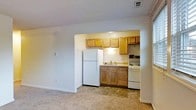 This single-floor residential unit offers a practical and straightforward layout designed for efficient everyday living. The space features an open-concept design where the living area flows seamlessly into the kitchen nook, creating a cohesive and functional environment. The kitchen is outfitted with warm honey-toned wood cabinetry both above and below the counter, providing ample storage space. Essential appliances are included, among them a full-size refrigerator, an electric range, and a built-in range hood, all finished in white for a clean, uniform look. The kitchen also features a single-basin sink with under-counter cabinetry and a tile floor, adding a practical, easy-to-clean surface in the cooking area. The adjoining living space is finished with neutral-toned carpeting, contributing to a cozy atmosphere. The walls throughout are painted in a soft, neutral white, keeping the space bright and versatile. Overhead, a ceiling vent ensures consistent climate control throughout the unit. The overall ambiance is modest and functional, making this an accessible and well-equipped space suited for comfortable day-to-day living.