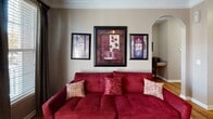 This single-floor residence offers a thoughtfully designed layout with warm, cohesive interiors throughout. Rich hardwood flooring runs through the main living areas, complemented by crown molding that adds a refined architectural touch. The entryway opens directly into the living space, where a bold red sofa anchors the room beneath a curated gallery wall arrangement, with an arched architectural detail providing an elegant transition between areas. Natural light filters in through a window dressed with dark curtain panels, adding warmth to the space. The kitchen features light wood cabinetry, a contrasting dark refrigerator, and a peninsula with granite-style countertops that doubles as a casual dining surface, seamlessly connecting to a dedicated dining area. The bedroom is a restful retreat with plush carpeting, warm taupe walls, and framed artwork above the bed. A walk-in closet is accessible directly from the bedroom, providing generous storage. The bathroom features a wide granite-topped vanity with an undermount sink, large mirror with overhead lighting, and ample counter space. Throughout the residence, a consistent warm color palette of taupe, burgundy, and gold tones creates a cohesive and inviting ambiance, making this a comfortable and functional living space.