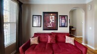 This single-floor residence offers a thoughtfully designed layout with warm, cohesive interiors throughout. Rich hardwood flooring runs through the main living areas, complemented by crown molding that adds a refined architectural touch. The entryway opens directly into the living space, where a bold red sofa anchors the room beneath a curated gallery wall arrangement, with an arched architectural detail providing an elegant transition between areas. Natural light filters in through a window dressed with dark curtain panels, adding warmth to the space. The kitchen features light wood cabinetry, a contrasting dark refrigerator, and a peninsula with granite-style countertops that doubles as a casual dining surface, seamlessly connecting to a dedicated dining area. The bedroom is a restful retreat with plush carpeting, warm taupe walls, and framed artwork above the bed. A walk-in closet is accessible directly from the bedroom, providing generous storage. The bathroom features a wide granite-topped vanity with an undermount sink, large mirror with overhead lighting, and ample counter space. Throughout the residence, a consistent warm color palette of taupe, burgundy, and gold tones creates a cohesive and inviting ambiance, making this a comfortable and functional living space.